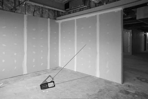 Micro Laser Distance Meter With Bluetooth Sync And Tilt Sensor placed in unfinished room, measuring angles with a clear backlit screen.