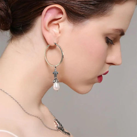 Woman wearing large hoop earrings with ornate design and dangling pearl.