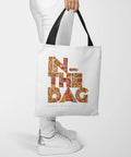 Person holding a tote bag with colorful "IN THE BAG" text design, wearing white pants and sneakers.