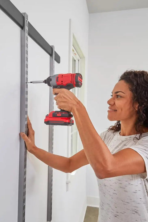 Woman assembling customizable wall shelving system with adjustable hooks and heavy duty rails using a power drill.