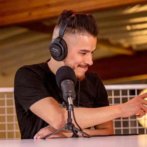 Man in headphones at podcast table with dynamic mic, using The Ultimate Two Host Podcast Stack Featuring Multitrack Recorder And Dynamic Mics Paired With Headphones Shielded Xlr Cables Tripod Stands And Foam Windscreens For Streaming That Sounds Gnarly And Helps You Drop Bangers On Daily Grind.
