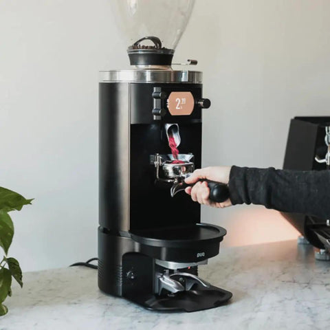 Automatic Espresso Tamper Knocks Out Consistent Pucks Every Single Time With Adjustable Pressure Settings Perfectly Calibrated For Mahlkonig E80 Grinders Smooth Operation In A Sleek Black Housing 58 Millimeter Size, demonstrating use beneath grinder.