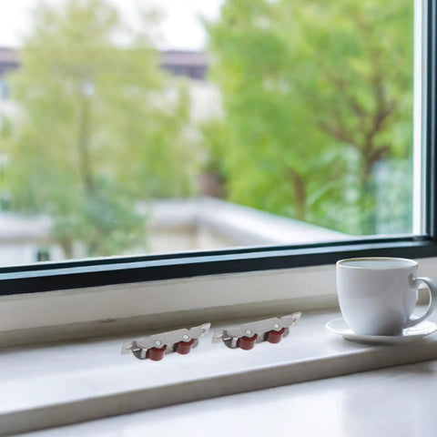 Wheeled Stainless Steel Sliding Window Rollers Feature Whisper Quiet Ball Bearing Action And Dead Smooth Gliding Motion Includes Four Heavy Duty Track Pulleys That Will Not Quit displayed on a windowsill with a cup of coffee.