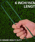 Hand holding Heavy Duty Green Landscape Staples 50 Pack 6 Inch Garden Stakes For Turf And Ground Fabric 11 Gauge Galvanized Lawn Pins Ideal For Weed Barrier Sod Fences Irrigation Tubing And Outdoor DIY Projects over a green background.