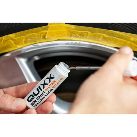 Hand holding QUUIXX rim paint bottle preparing to touch up black rim as part of Solid Alloy Wheel Repair Kit For Black Rims Smooths Out Curb Rash And Road Scuffs DIY Two Part Liquid Aluminum Compound Plus Spot On Black Touch Up Paint For A Fresh New Ride Glow Up.