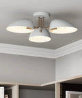 Modern flush mount ceiling light with three white shades and brass accents on a ceiling above bookshelves.