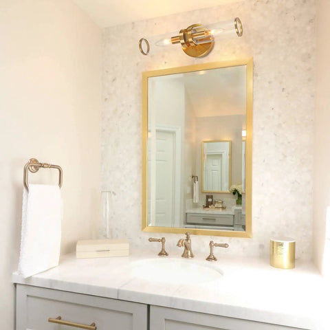 Bathroom with gold wall sconce lights two pack with candle shaped glass shades above mirror, brass fixtures, and marble countertop.