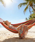 Woman relaxing in Rainbow Cotton Canvas Hammock With Tree Straps And Bar, Ultra Comfy Durable Fabric on a beach between palm trees.
