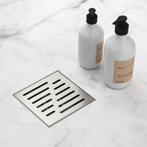 6 Inch Square Shower Drain With Brushed Nickel Surface Premium 304 Stainless Steel Build Features Smart Hair Catcher on marble floor with bottles.