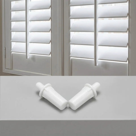 Two white spring loaded shutter pins for plantation window shades on a gray surface with window blinds in the background.