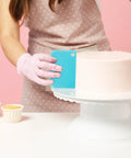Next Level Cake Decorating Kit Featuring Hella Durable Anti Burst Piping Bags Stainless Steel Frosting Tips Set And Reusable Pastry Supplies For Baking Like A Champ Zero Worries, featuring a cake being smoothed with a blue scraper.