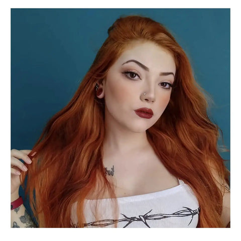 Portrait of a woman with long orange hair, wearing a white top, showcasing a nose stud and piercings against a blue background.