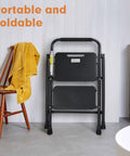 Heavy Duty 2 Step Folding Ladder With 500 Lb Capacity Anti Slip Wide Pedals in black, folded, in home setting next to chair.