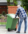 Person moving green garbage can with black wheels, demonstrating Garbage Can Wheels Replacement With Metal Hollow Shaft And Small Rubber Wheels 19.68 X 7.28 X 3.93 Inches Quiet Trash Can Pulley For Easy Installation And Efficient Waste Collection in a garden setting.