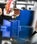 Close-up of a hand using tweezers to insert a cleaning needle into a 3D printer nozzle, part of 3D Printer Nozzle Cleaning Kit 13 Piece Set.
