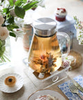 One-Touch 2-In-1 Tea Maker Teapot and Kettle with stainless steel filter lid, filled with tea, on a table setting with flowers.