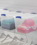 Twenty Seven Quart Clear Plastic Storage Container With Durable Blue Latches And Secure Recessed Lid Stackable Tote Bin For Hassle Free Home Organization storing folded clothes on a wire shelf.