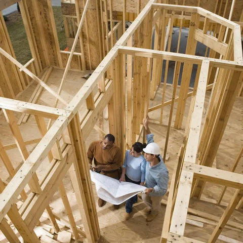 Three people reviewing construction plans inside a wooden house framework under construction.