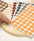 Heavy Duty Self Adhesive Screw Hole Caps 540 Pack Slick Yellow Wood Grain Finish on sheets with finger pointing to cap.