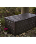Lockable 120 Gallon Outdoor Storage Deck Box for Patio Backyard and Garden in Brown Waterproof Plastic Bench among plants.