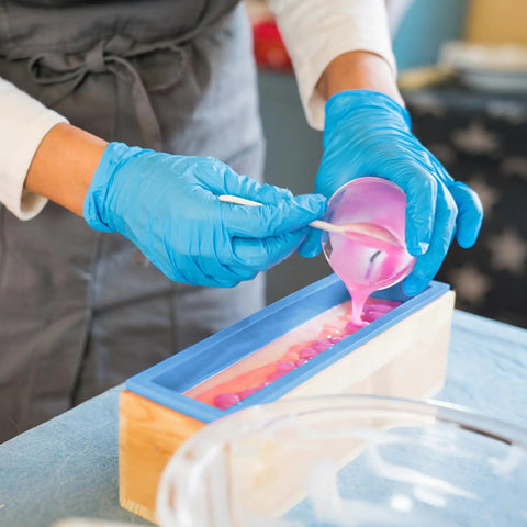 Person pouring pink soap mixture into Ultimate 7 Piece Soap Making Kit With Silicone Molds And Stainless Steel Slicers, wood box visible