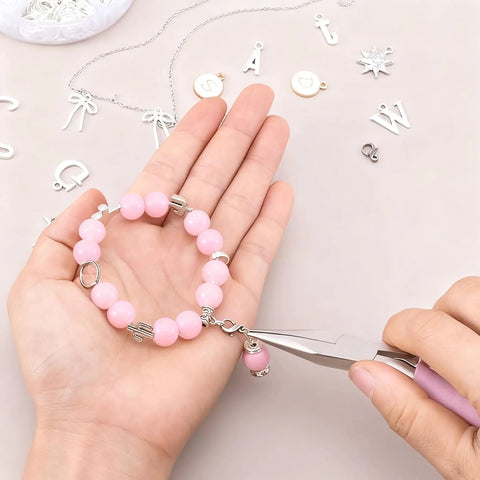Person using pliers to attach a pink beaded bracelet with silver charms, surrounded by silver jewelry components on a table.