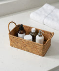 Spacious Open Top Seagrass Woven Storage Basket With Heavy Duty Handles For Kitchen Farmhouse Pantry Bathroom Towels Cosmetics And Hassle Free Daily Organization Goals on a countertop with toiletries.