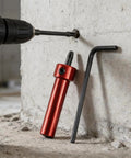Side view of a red metal socket adapter with hex drive, next to an L-shaped hex key and drill on concrete.