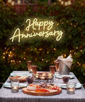 Outdoor anniversary dinner setup with Happy Anniversary LED neon sign and candle-lit table, evening garden ambiance.