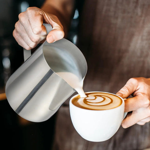 Four Pack 20 Oz Stainless Steel Milk Frothing Pitchers For Coffee And Latte Art Barista Gear Steamer Jugs Cappuccino Maker Pro Grade Stainless Steel Home Cafe And Shop Essentials being used to pour milk into a cappuccino cup with latte art.