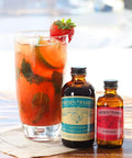Glass of iced beverage with cucumber and strawberry garnish next to Nielsen-Massey Tahitian vanilla and rose water bottles.