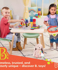 Two toddlers playing with colorful B. toys playset at a table with stuffed bunny on carpeted floor and window curtains in background.