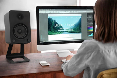 Rock Solid Elevated Desktop Speaker Stands For Large Bookshelf Speakers And Full Size Studio Monitors Six Point Nine Inch Tall Twenty Two Lbs Reduces Resonance With Two Point Five Millimeter Damping Padding One Quarter Twenty Mounting Pair Black next to computer on desk with person
