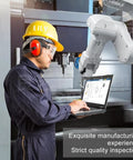 Worker using a laptop to monitor a robotic arm in a manufacturing setting with a yellow safety helmet.