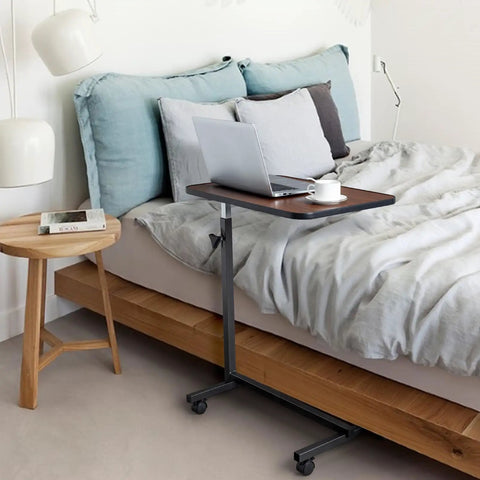 Smooth Rollin Mobile Adjustable Height Portable Computer Desk With Lockable Casters For Top Tier Home Office Standing Laptop Workstation Dark Wood Finish beside a bed, with laptop and cup.