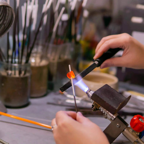 Hands using a torch to heat glass rod in crafting process, surrounded by tools and equipment on a workbench.