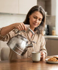 Woman pouring hot water from a Whistling Tea Kettle For Stovetop 2.3 Quart Stainless Steel Teapot into a mug in the kitchen.