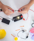 Hands connecting E10 Screw Base Miniature Bulbs For DC 12V Projects Packing Ten Warm Yellow Glow to a battery with clips.