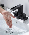 4 Inch Centerset Vanity Sink Faucet With Dual Handle Waterfall Spout Three Hole Basin Mixer Dope Matte Black Finish With Overflow Pop Up Drain Easy Install Bathroom Tap For Modern Bathrooms with water flowing over hand.