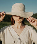 Woman in a sun hat holding the brim, with a flower in her mouth, against a serene green field backdrop.