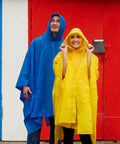 Two adults in blue and yellow ponchos smiling against a colorful wall. Lightweight Hooded Packable Poncho With Side Snaps For Adults Travel Ready Rain Gear That Packs Into Its Pouch Reusable Water Resistant Rain Shield To Keep You Dry In Any Forecast.