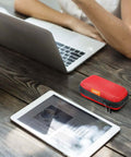 Small Red Rectangular Eva Case With Hella Tough Crush Proof Hard Shell And Smooth Zipper Mesh Pouch For Charger Cables Earbuds Memory Cards And Everyday Carry on a wooden desk near a laptop and tablet.