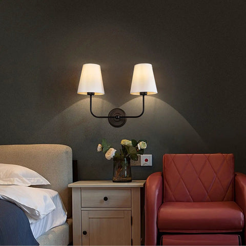 Double Light Wall Sconce With White Fabric Tapered Shades In Black Finish For Your Bathroom Vanity Hallway Stairway And Living Room Two Light Fixture With E26 Socket Dimmable LED Compatible displayed in a bedroom setting above nightstand and chair.