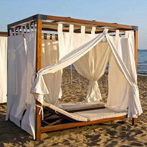 Wooden pergola with white curtains on a beach, featuring a lounge bed inside, set against an ocean background.