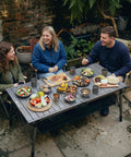 People enjoying a meal on a 50 Inch Camping Table With Adjustable Height Legs For Uneven Ground, Easy Setup Portable Folding Aluminum Roll-Up Table With Mesh Pocket For Camping, Picnic, BBQ, Beach And Outdoor Cooking.