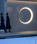 Two women walking past a massive 80cm Outdoor Wall Light Brushed Stainless Steel Round LED Sconce on a modern wall.