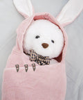 Stuffed animal wrapped in pink bunny-ear blanket with a plaid bow tie, fastened with clips, on a gray background.
