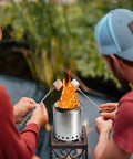 Two people roasting marshmallows over a Pellet Tabletop Fire Pit 7 Inch Mini Outdoor Fire Pit With Removable Ash Pan Carry Bag Marshmallow Roasting Sticks Loading Spoon Ideal For Patio Camping Backyard Picnics on a patio.