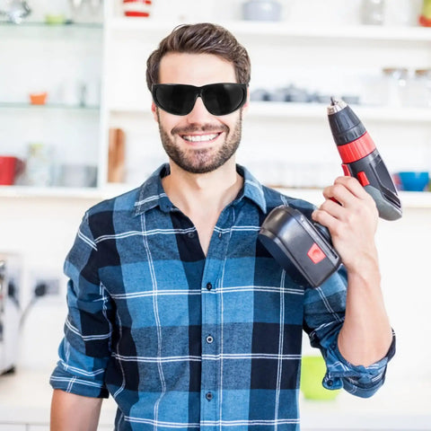Welding Safety Goggles Offering A Badass Level Of Protection With An Over Glasses Design Delivering Crystal Clear 180 Degree Vision So You Can See Every Detail With Ease And Comfort Using High Quality Materials To Keep You Safe And Secure During Work, worn by a man holding a cordless drill.