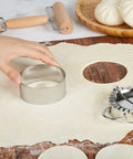 Hand using a stainless steel dough cutter on rolled dough for making dumplings for Real Deal 10 Inch Bamboo Steamer Basket Set.